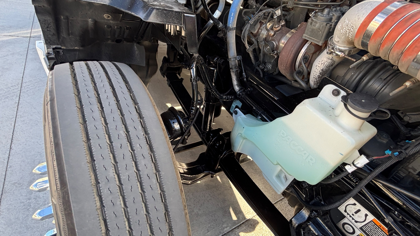 Kenworth T680 engine bay showing coolant reservoir with greenish fluid and various hoses; front tire tread visible and engine components in view.