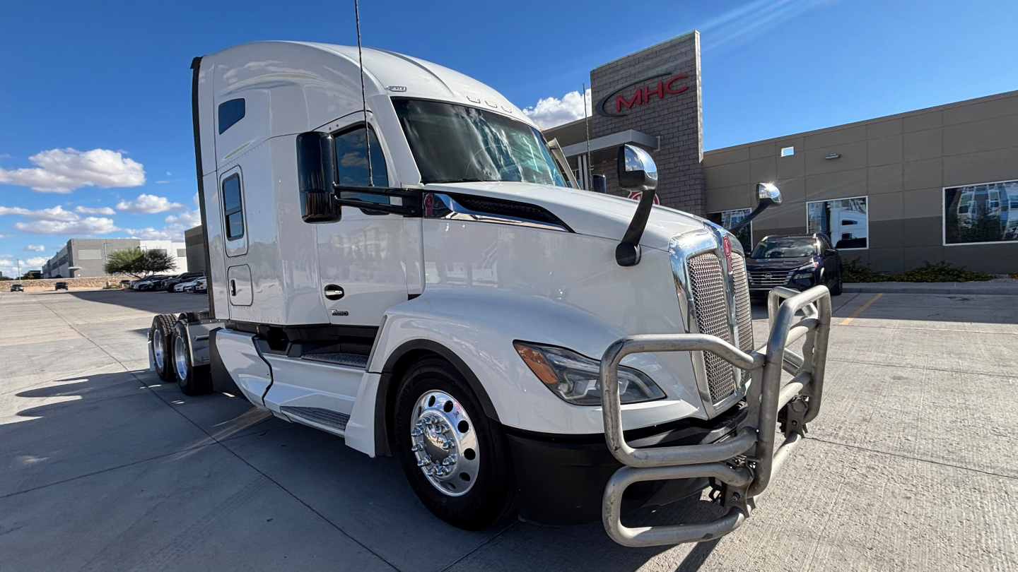 Kenworth T680 conventional sleeper truck in white showing front grille guard, dual exhaust, and side mirror; daytime dealership lot in background.