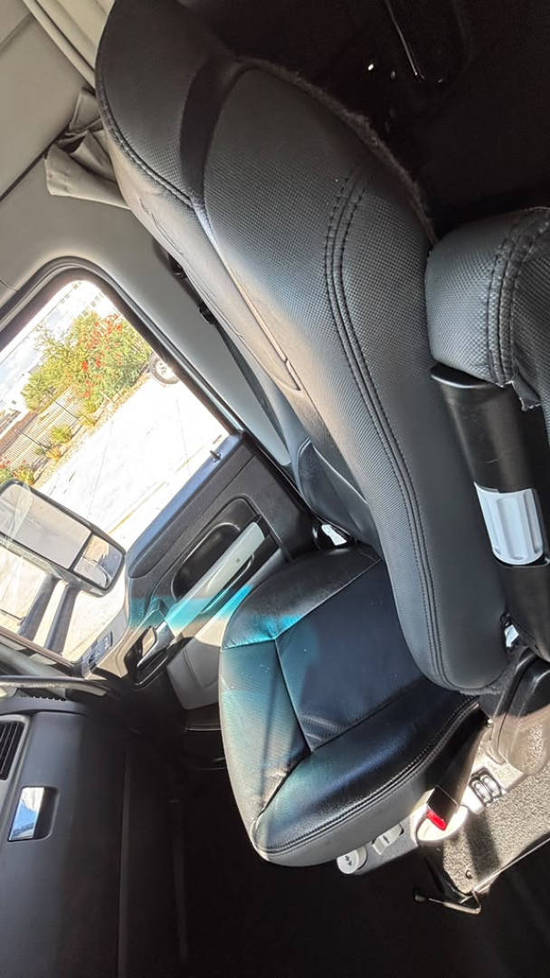 Kenworth T680 interior seat area with driver seat and partial dashboard; light grey and black seats, window and door panel visible.
