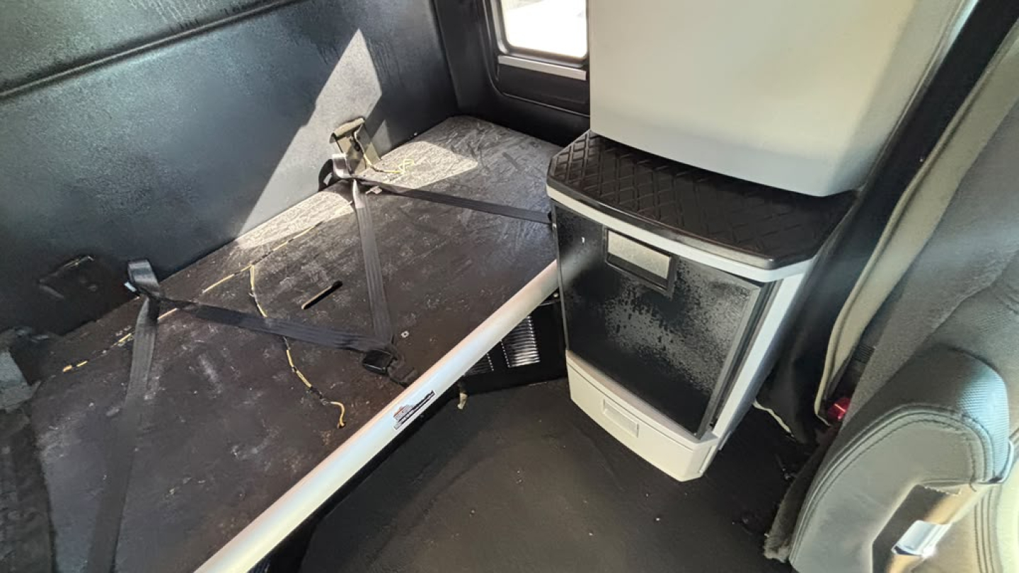 Kenworth T680 interior sleeper cab area with driver seat and overhead storage, showing a metal platform and seat belt anchors.