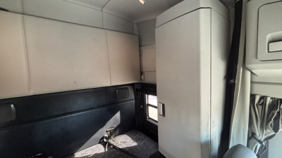 Kenworth T680 cab interior showing sleeper area with light gray cabinets and window; small bench area with black lower wall, light from side window.