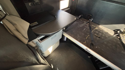 Kenworth T680 truck interior showing sleeper cab with flatbed/bed area and storage bin; seating area on left, utilitarian cabin with dark panels and light from window on right.