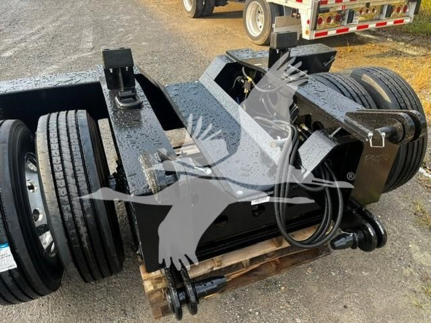 Trailer axles and flip axle assembly with winch and hydraulic lines visible. Metallic frame with black components, dual wheels, and a coiled hydraulic hose lying on the frame.
