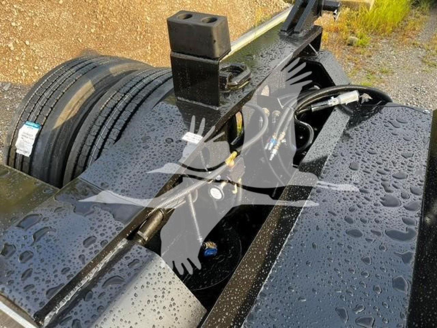 Fontaine Flip Axle trailer component view showing hydraulic hoses and mounting frame with wet surface and spare tire in background.