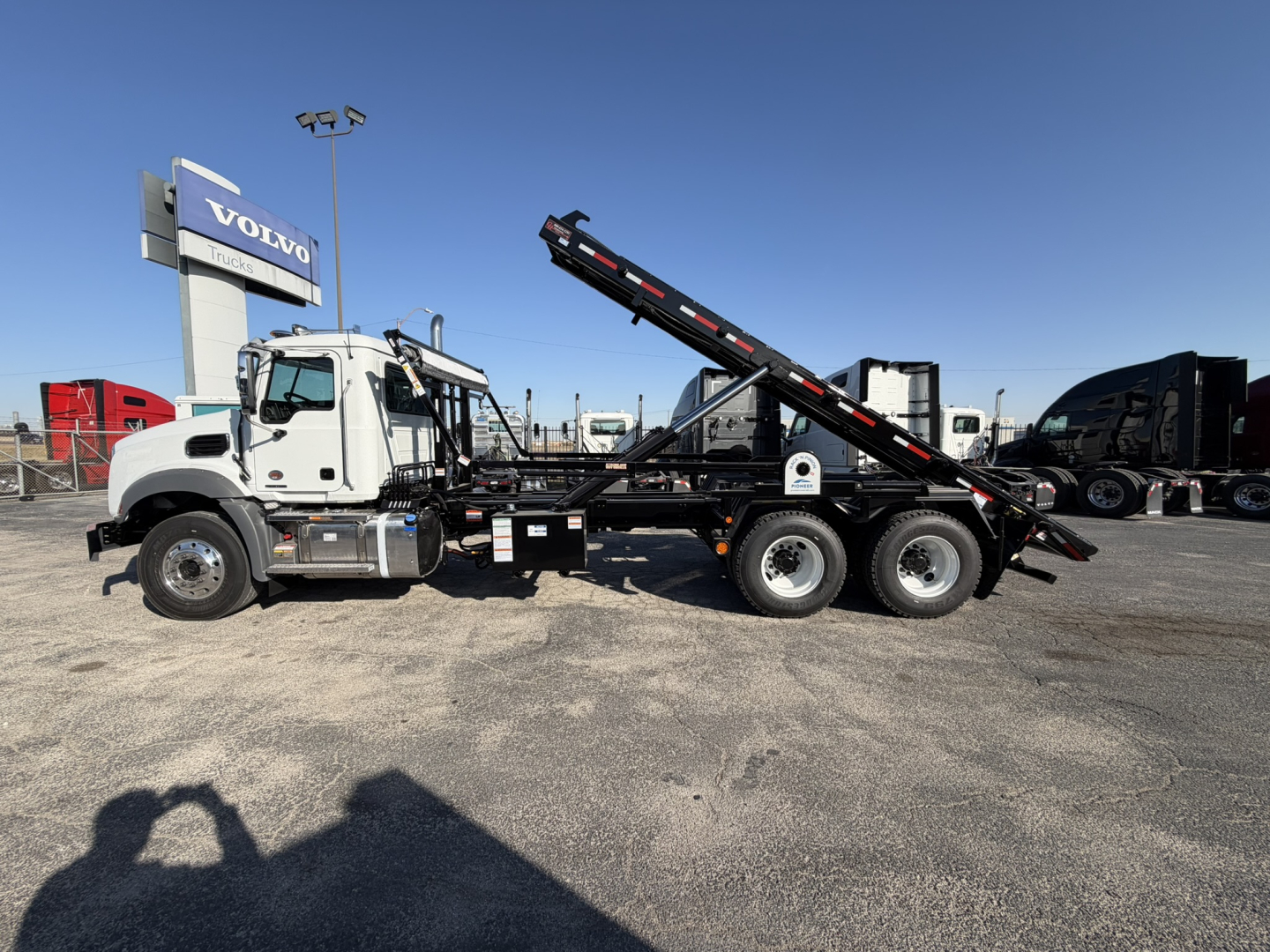 Truck side view showing Mack GR64B roll-off truck with hydraulic loading system and black tilt-bed in front of Volvo sign.