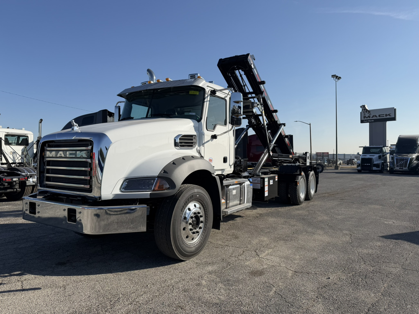 Truck shows a white 2026 Mack GR64B roll-off, side view with loading ramps in raised position and chrome bumper.