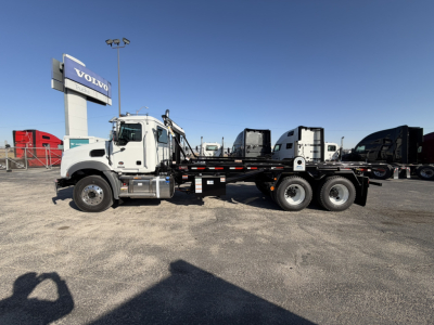 2026 Mack GR64B rolloff truck side view showing cab, long flatbed, and dual rear axles with a white cab and black chassis.