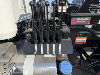 Hydraulic lever controls on Mack GR64B roll-off truck showing control levers with white labels and blue coolant cap nearby; warning tag and dealership sticker visible in background.