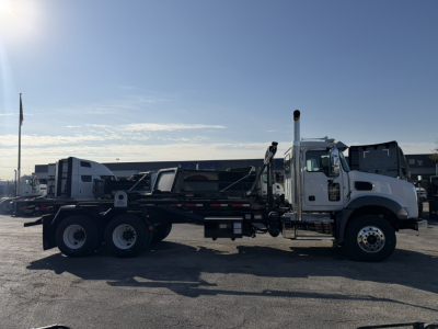 2026 Mack GR64B roll-off truck with flatbed, side view showing dual rear axles and hoist mechanism.