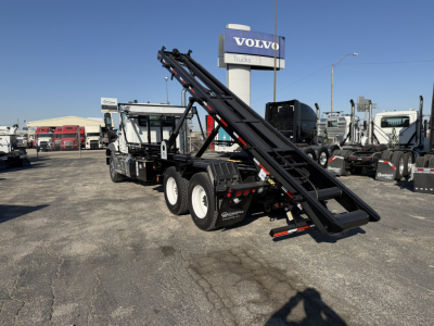 Mack GR64B roll-off truck with inclined loading ramps in a dealership lot, black and white cab, Volvo Trucks sign in background.