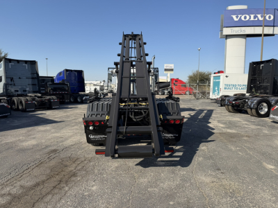 View of a 2026 Mack GR64B rolloff truck chassis from rear, with a tall black grapple/loader arm and hydraulic components elevated. In the background, a Volvo dealership sign and other trucks are visible.