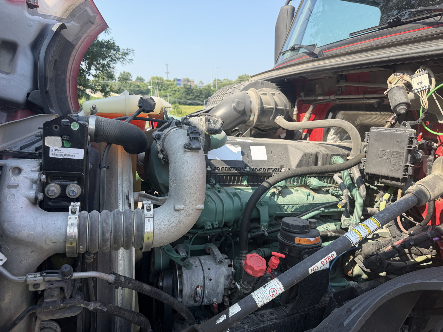 Truck engine bay showing green Volvo engine with hoses, belts, and components; red hood and surrounding frame visible.