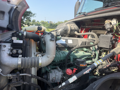 Truck engine bay showing green Volvo engine with hoses, belts, and components; red hood and surrounding frame visible.