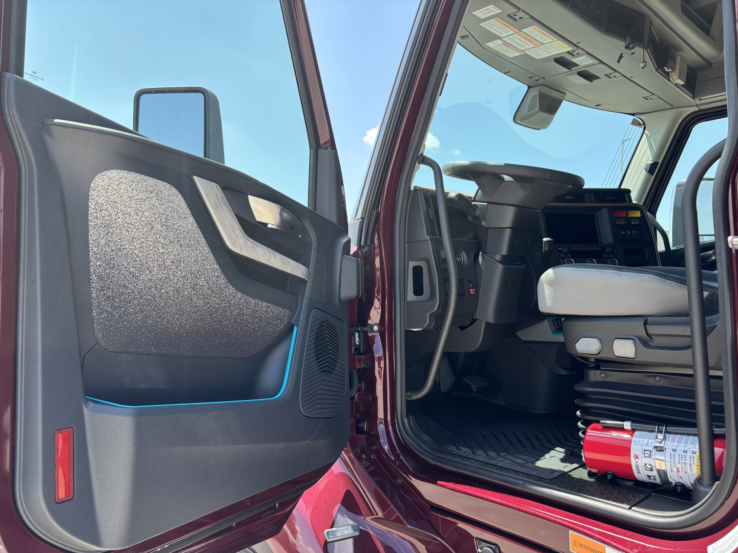 Truck cab interior the door open showing black interior with blue accents, dashboard, seating, and a red extinguisher on the floor near the seat.
