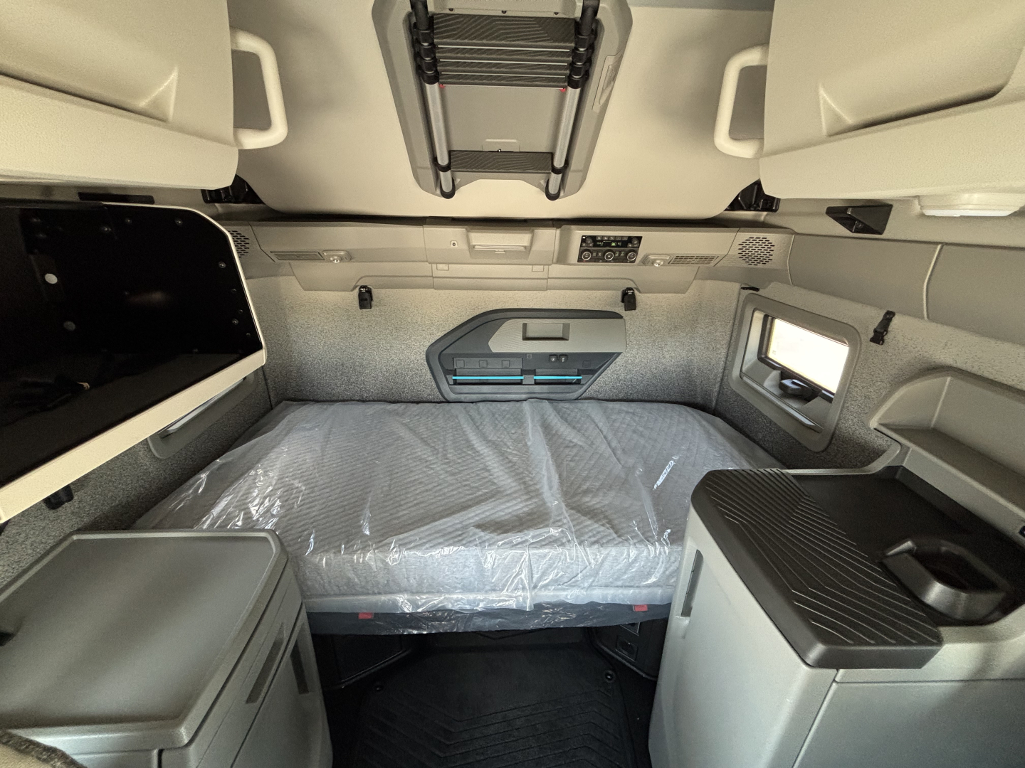 Volvo VNL64T860 sleeper cab interior showing bunk bed with plastic-covered mattress, overhead storage, scratchless panels, and side workspace in neutral tones.