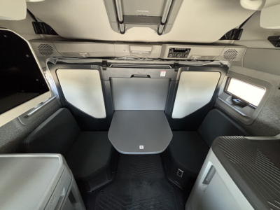 Interior of a Volvo VNL cabin with a table in the sleeper area, light gray panels, seating on both sides, overhead storage, and control panel on the front wall.