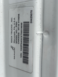 Mack Trucks label showing model, VIN, and serial numbers on a white surface; vertical orientation with barcode. Text includes 'MACK TRUCKS INC', 'MODEL DUMP', 'MODEL PI64', 'VIN: 1M2PN4GC6TM019698', 'STOCK: 70571', '187? 344-...'