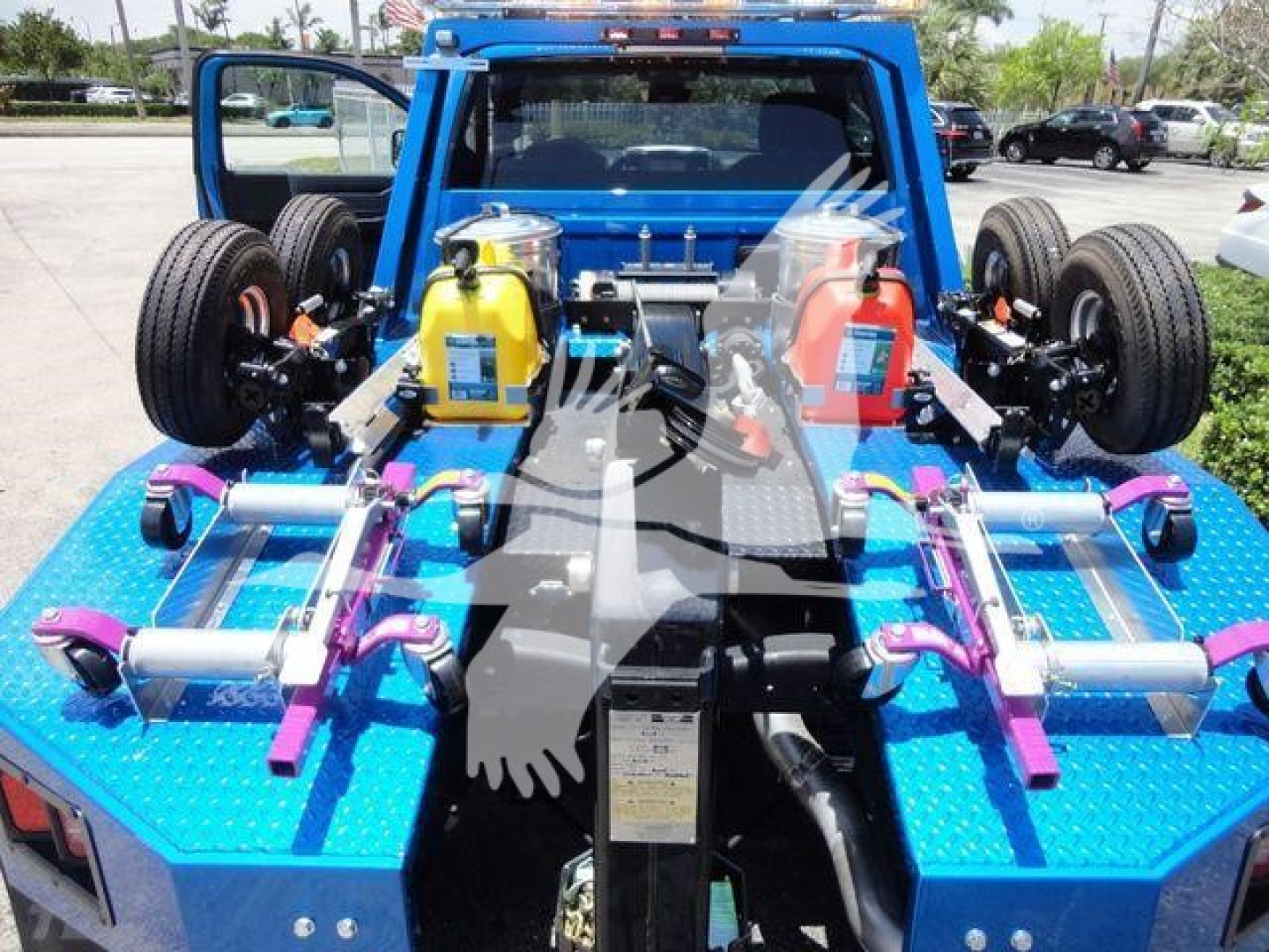 Vehicle rear showing dual frame rails and winch setup with yellow coolant reservoir and blue flatbed, two wheels visible, hydraulic and tie-down components.