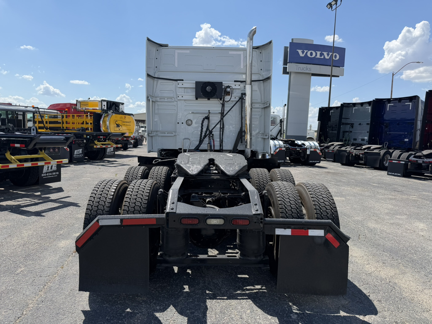 Truck rear view showing fifth wheel and rear chassis with mud flaps and taillights; Volvo dealership sign in background.