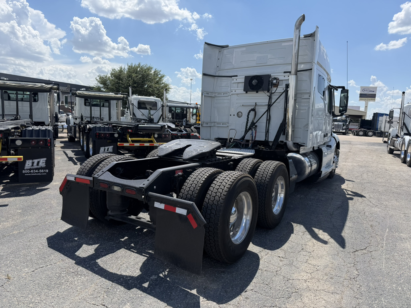 Truck chassis with cab in rear view, showing fifth wheel and dual rear axles, white sleeper cab, rear mud flaps, exposed wiring on back of cab, blue sky with clouds.