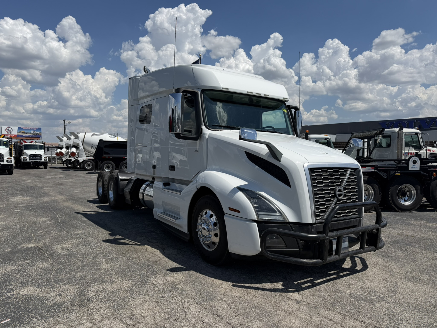 Volvo VNL64T740 sleeper tractor in white, front grille guard, large side mirrors, day cab, with other trucks in lot and blue sky.