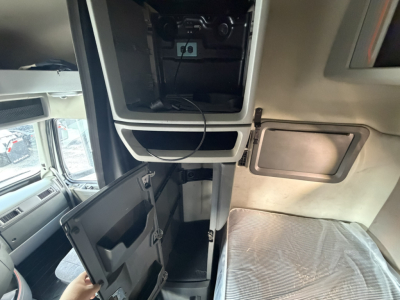 Volvo VNL64T740 sleeper cab interior showing overhead bunk compartments and open storage cabinet with cords, side door, and cockpit area.