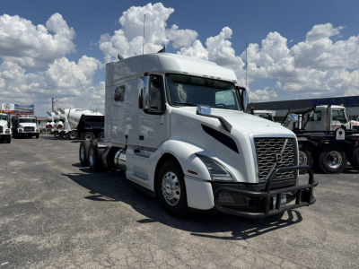 Volvo VNL64T740 sleeper tractor in white, front grille guard, large side mirrors, day cab, with other trucks in lot and blue sky.