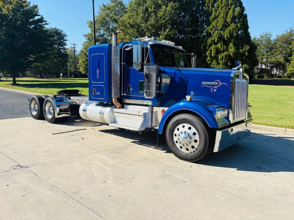 Kenworth W900L truck in blue profile view showing front grille, chrome bumper, side exhaust, and dual rear axles.