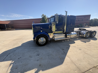 Kenworth W900L side view showing blue exterior cab and chrome fuel tanks, triple rear axles, and conventional sleeper configuration.