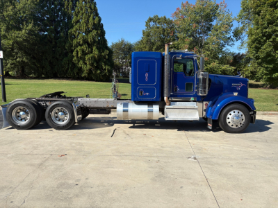 Kenworth W900L truck side view showing blue cab with chrome exhaust stack and fuel tank, extended chassis with triple rear axles.