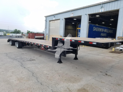 Flatbed trailer with dropdeck beavertail, showing a long low-profile deck and dual rear axles, parked outside a workshop.