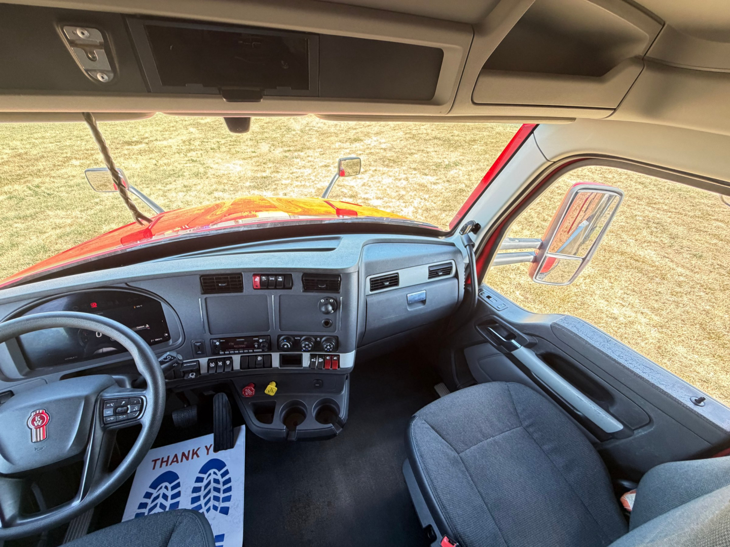 Cab interior of a Kenworth T680 truck showing steering wheel, dashboard with controls, and front seats; overhead compartment and sun visors visible in daylight.