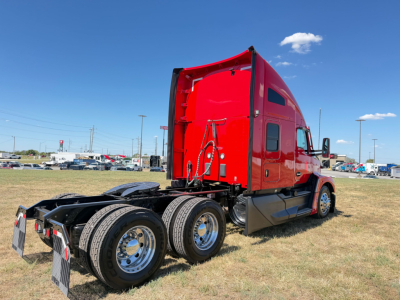 Kenworth T680 truck in red sleeper cab, rear-side view showing dual rear axles and chassis, all-terrain yard setting.