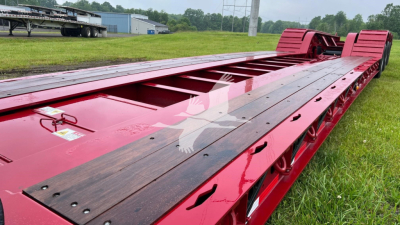 Flatbed trailer chassis showing red lowboy trailer with wooden deck planks and dual rear axle set, side rails, and ramps visible in distance.