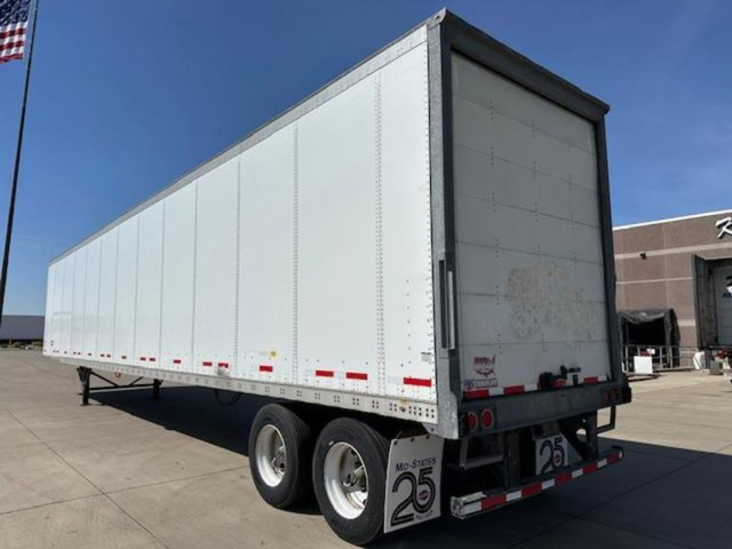 Trailer side and rear view showing a white enclosed van trailer with dual rear axles and minor dirt on rear doors. Visible are rear bumper, taillights, and reflective safety tape.