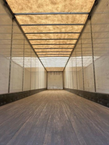 Trailer interior showing long cargo space with wooden floor and metal rafters, ceiling panels, and side walls.