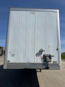 Rear View of a white 2016 Wabash van trailer showing door latch, power outlet, and label stickers. Visible text includes small manufacturer logos and QR sticker near left of center.