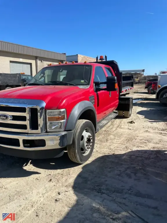 Ford F-550 crew cab dump truck in red with black bed sides, front grille and bumper, dual rear wheels, parked on a dirt lot with clear blue sky.