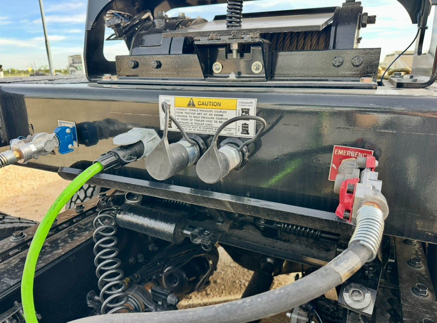 Chassis rear of a Landoll trailer showing hydraulic connections and warning decal, emergency red handle, hoses, and coil springs.