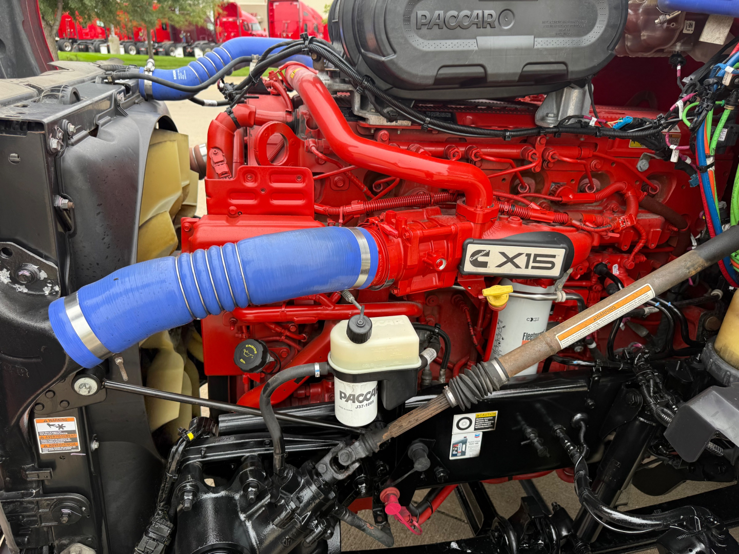 Kenworth T680 engine bay showing X15 badge, red engine components, blue hoses, coolant reservoir, and various hoses and wiring.