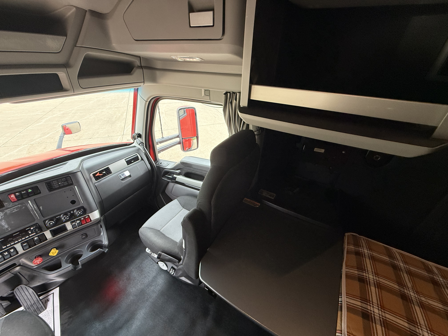 Interior cab area of a 2023 Kenworth T680 showing driver seat, dashboard, and bunk with overhead cabinet and plaid mattress cover.