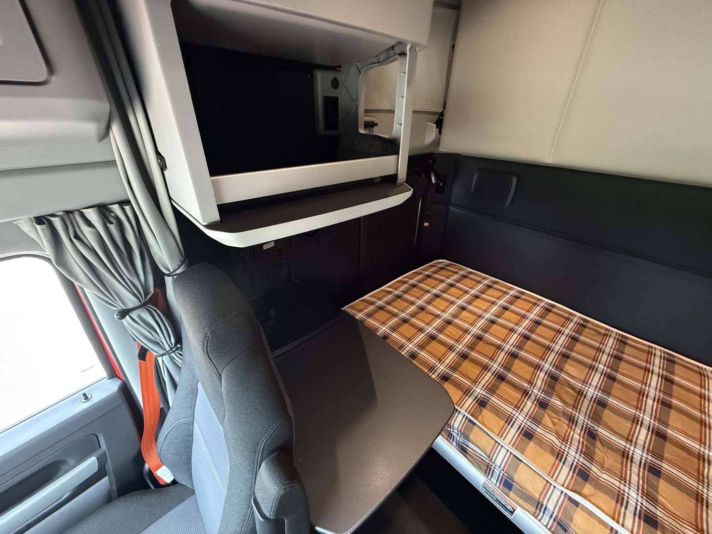 Interior of a Kenworth truck sleeper with a plaid blanket on the bunk and overhead storage shelf open.