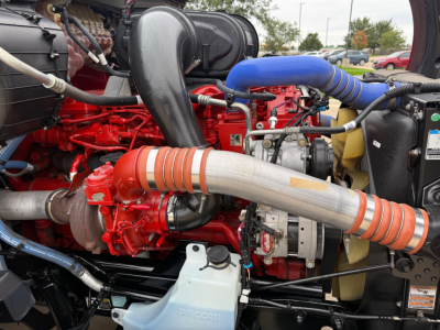 Kenworth T680 engine bay showing red engine block, large silver exhaust pipe, blue intake hose, and various hoses and components.