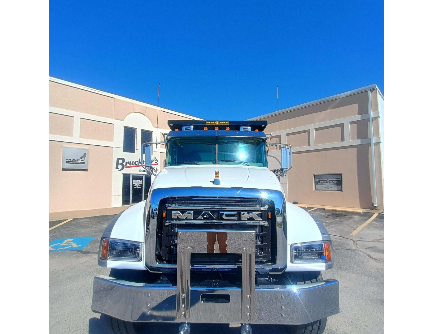 Truck front view showing Mack grille with bold MACK letters, chrome bumper, white cab, and blue sky; no readable model text beyond the brand name on grille.