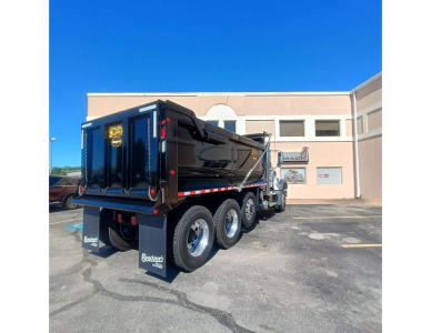 Dump truck rear and cab showing Mack GR84F dump truck with black dump bed and multiple axles; visible text includes 'MACK' on the bed, and 'BLACKHAWK' or brand sticker; no other legible labels.