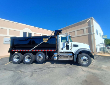 Truck side view of a white Mack GR84F dump truck in a lot, showing the dump bed, chrome exhaust, six wheels on the rear, and black trim.