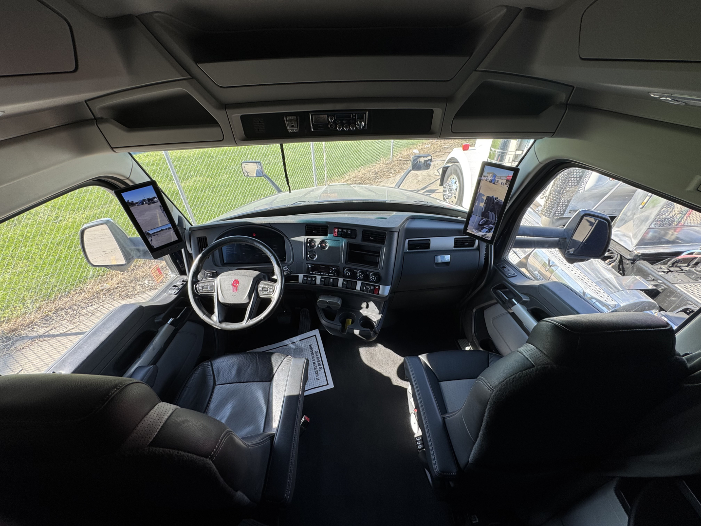 Kenworth T680 cab interior showing steering wheel, dashboard, two passenger screens, and driver/passenger seats.