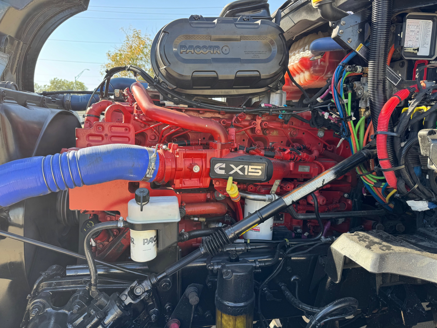 Kenworth T680 engine bay showing red Cummins engine with blue silicone hose and black air intake housing labeled Paccar MX-15 engine tag.