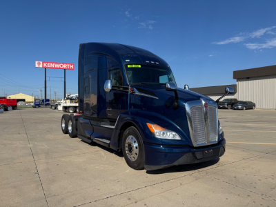 Kenworth T680 tractor in blue, front three-quarter view, highway tractor with sleeper cab, dealership lot in background.