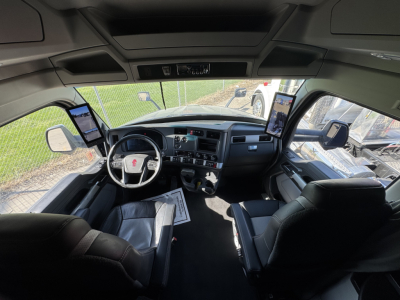 Kenworth T680 cab interior showing steering wheel, dashboard, two passenger screens, and driver/passenger seats.